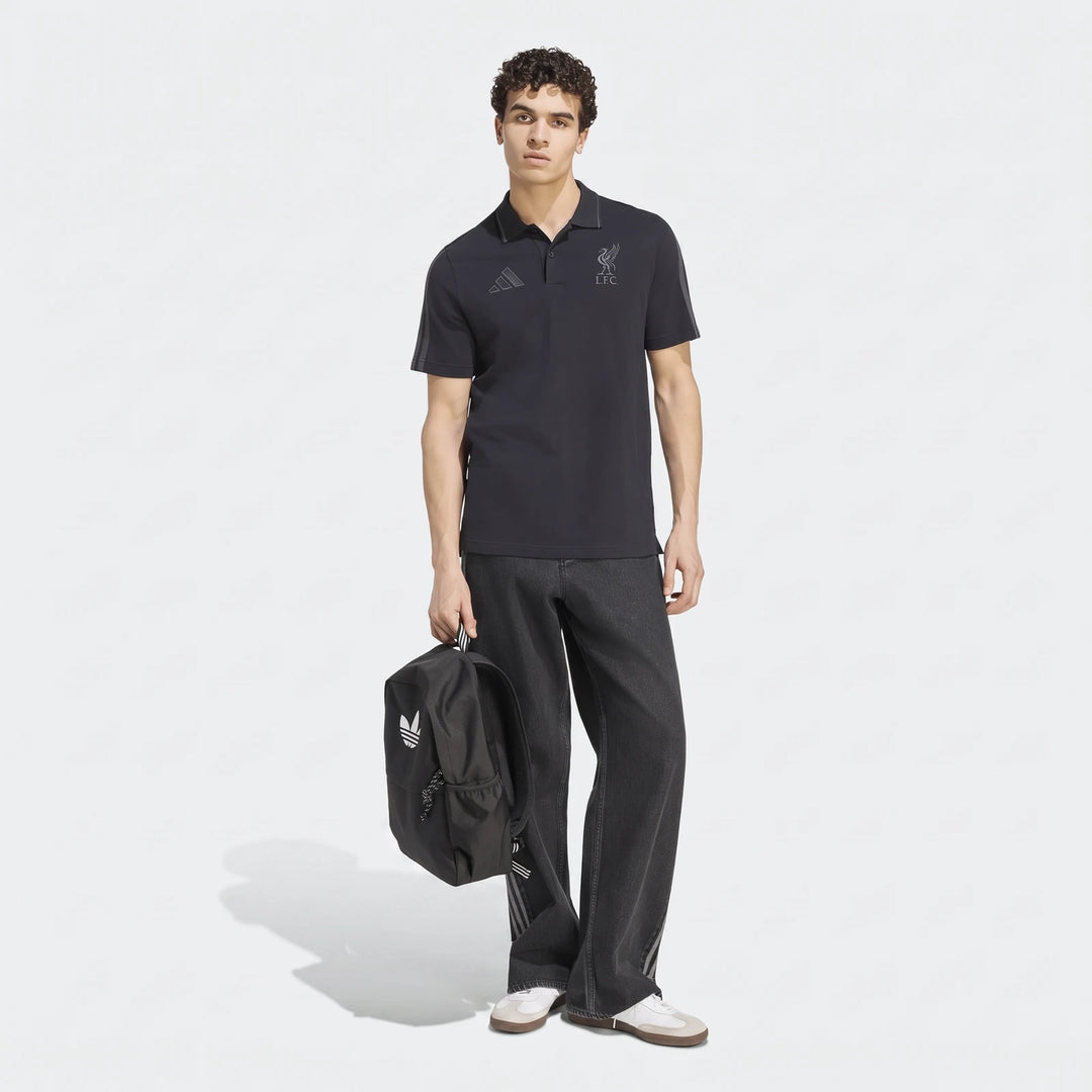 Male model wearing a black Adidas Liverpool FC DNA polo shirt, dark gray wide-leg jeans, and sneakers, holding a black