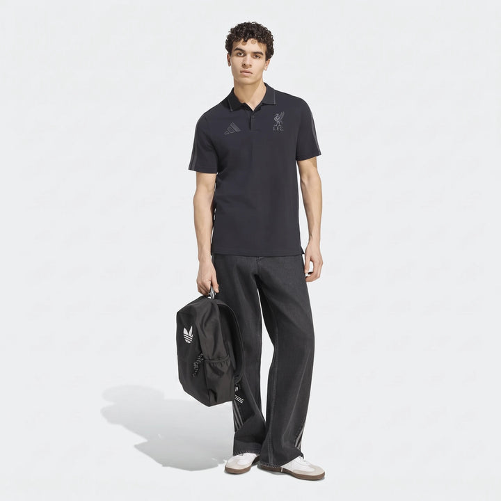Male model wearing a black Adidas Liverpool FC DNA polo shirt, dark gray wide-leg jeans, and sneakers, holding a black