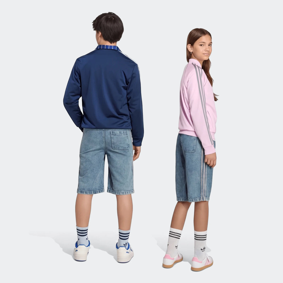 Two models, a boy and a girl, wear light wash loose denim jorts and track jackets with stripe details. Full body shot.