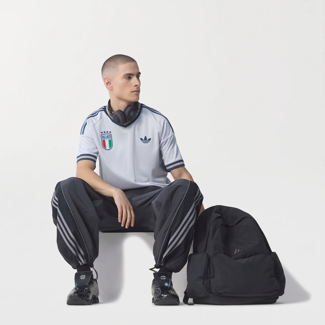 Male model wearing a light gray Adidas Italy football jersey, dark track pants, and black headphones, with a black backpack.