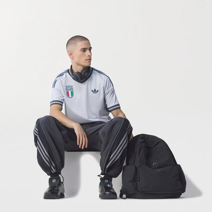 Male model wearing a light gray Adidas Italy football jersey, dark track pants, and black headphones, with a black backpack.