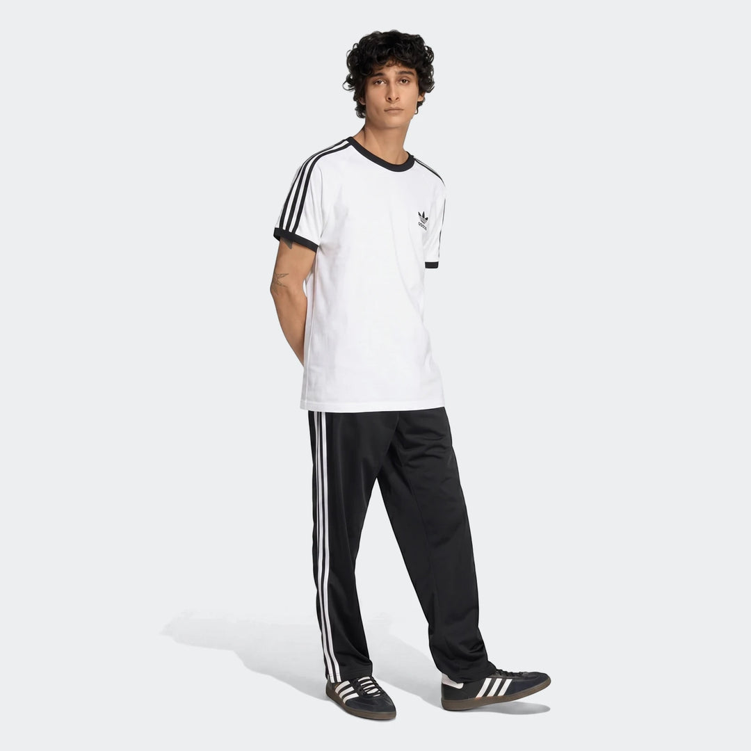 Male model wearing black Adidas Firebird Track Pants with white stripes, a white t-shirt, and black sneakers.