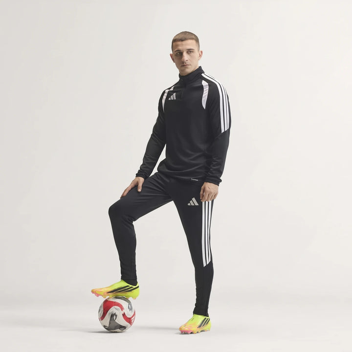Male model in black Adidas tracksuit and fluorescent F50 Hyperfast Elite Laceless Firm Ground Boots, with foot on soccer
