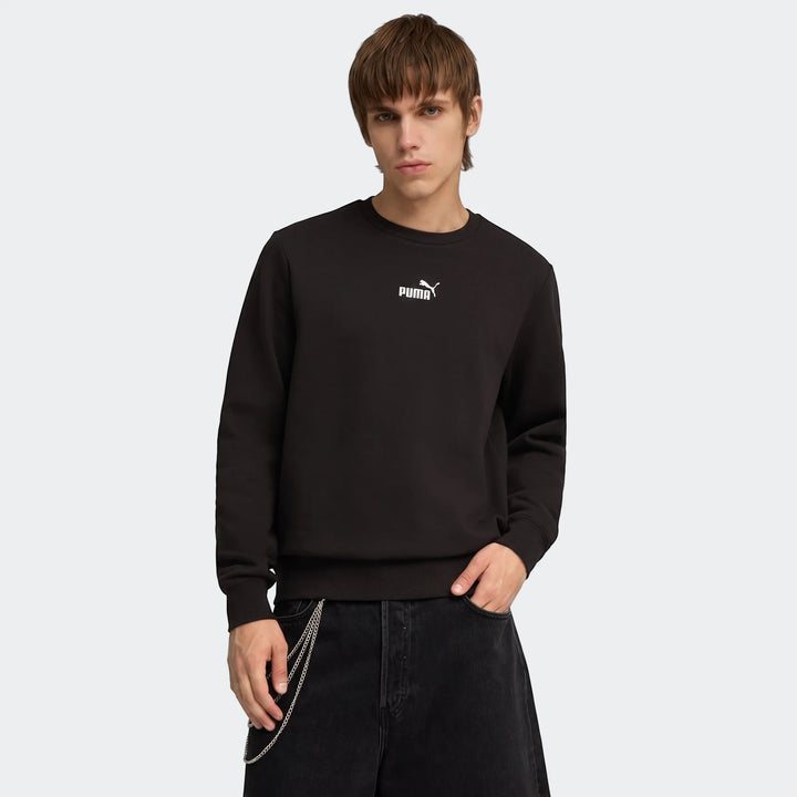 A male model wears a black Puma Ess No.1 Logo Crew sweatshirt, styled with black jeans and a silver chain, captured from the