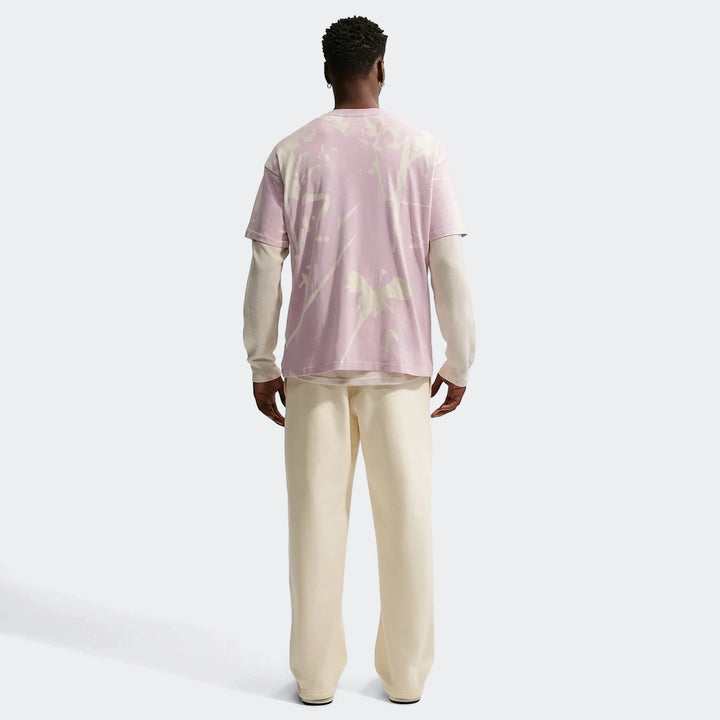 Rear view of a male model wearing a lilac tie-dye t-shirt over a cream long-sleeve shirt and cream sweatpants, standing on a
