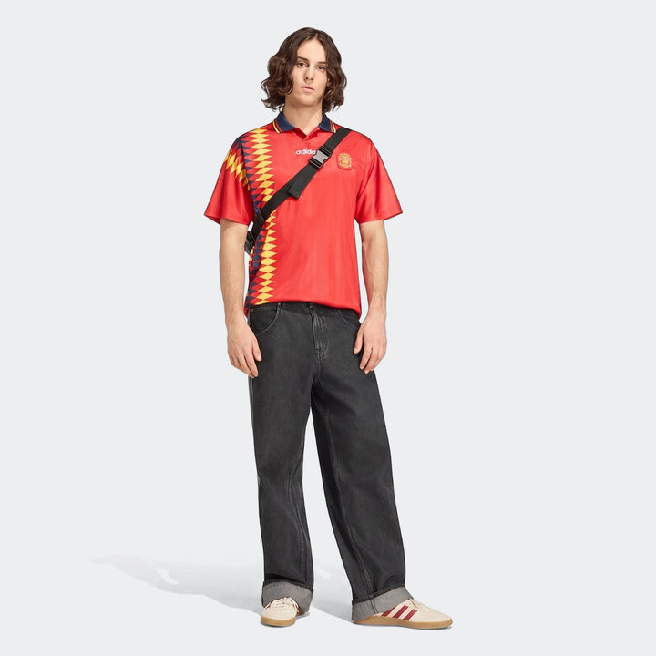 Male model wearing a red, yellow, and blue patterned Adidas soccer jersey, dark grey wide-leg jeans, and cream sneakers.