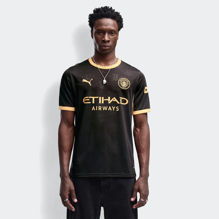 Manchester City 26/27 Away Jersey Men in black with gold accents, featuring bee pattern and team crest, worn by a male model.