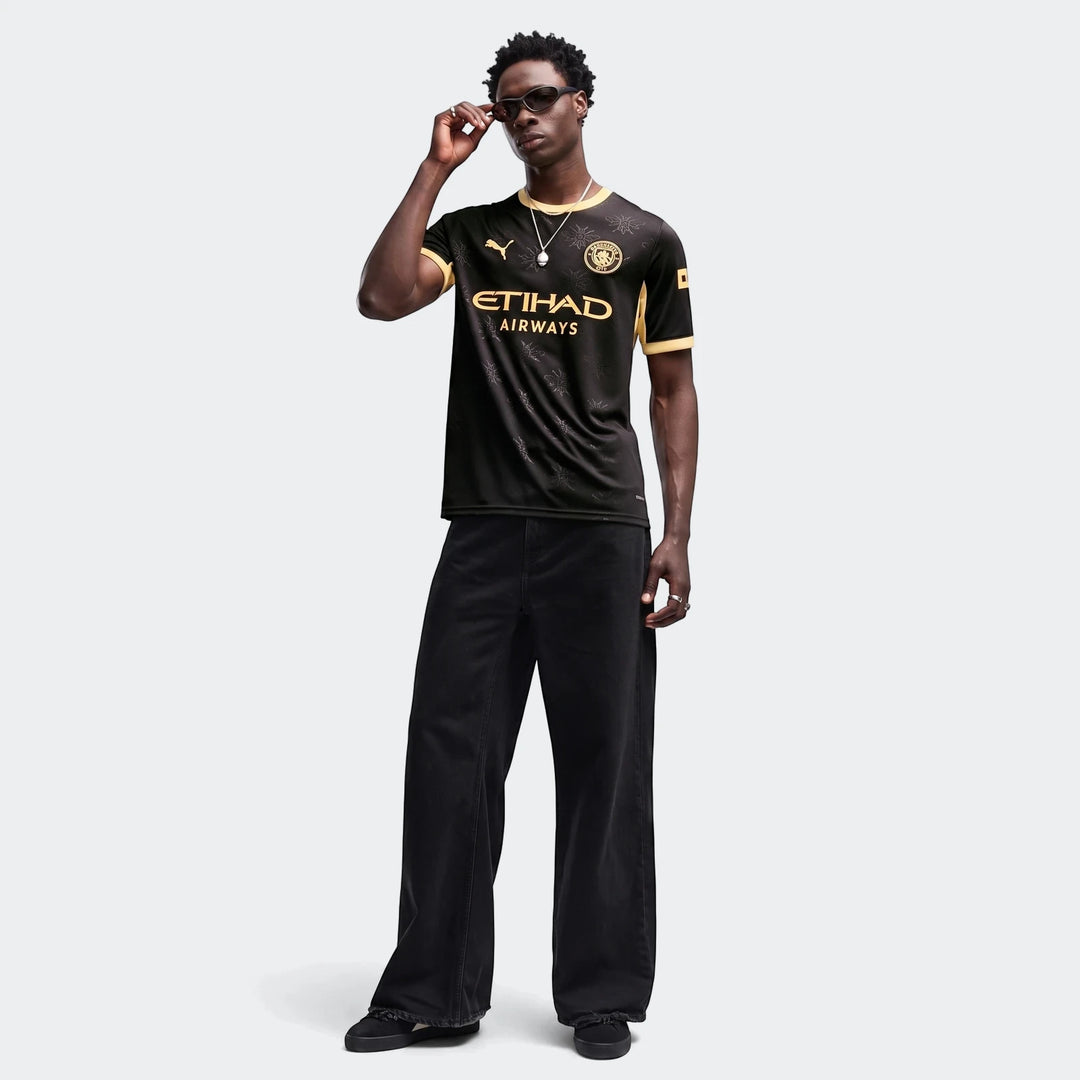 Male model wearing a black and gold Manchester City 26/27 Away Jersey with black pants and sunglasses, full body shot.