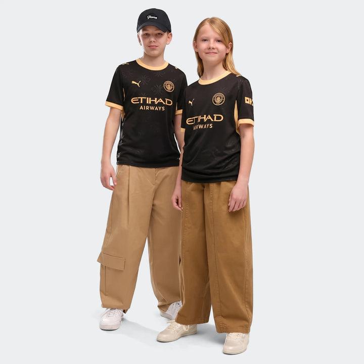 Two children, a boy and a girl, model the black and gold Manchester City 26/27 Away Youth Jersey, paired with tan pants.