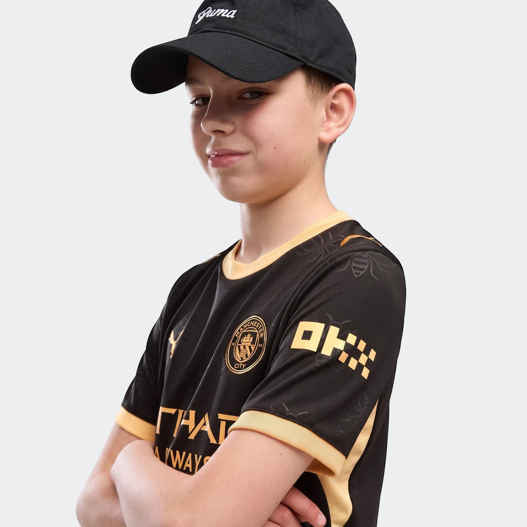 Young boy wearing a black and gold Manchester City 26/27 Away Jersey and a black Puma cap, arms crossed.