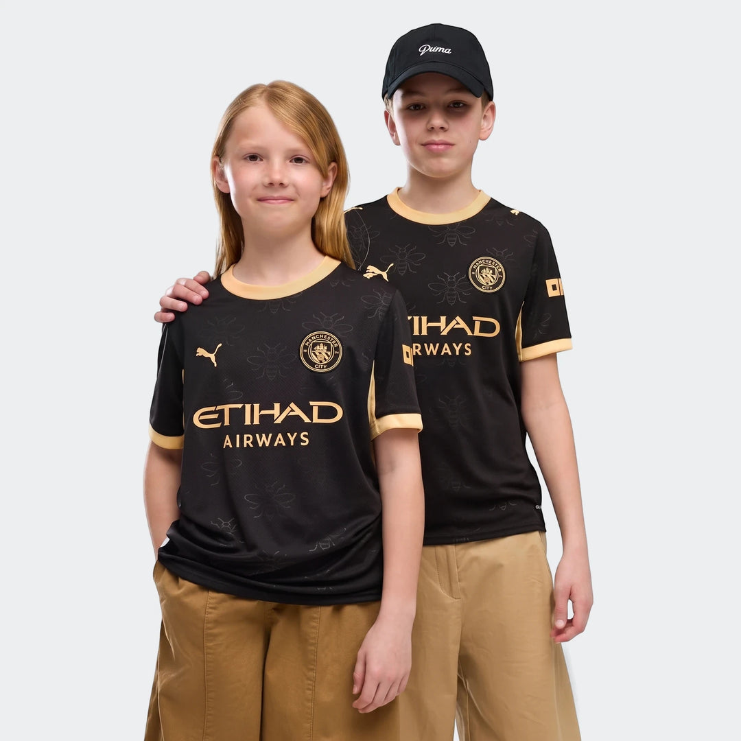 Two children model the black and gold Manchester City 26/27 Away Youth Jersey, featuring club crests and sponsor logos.