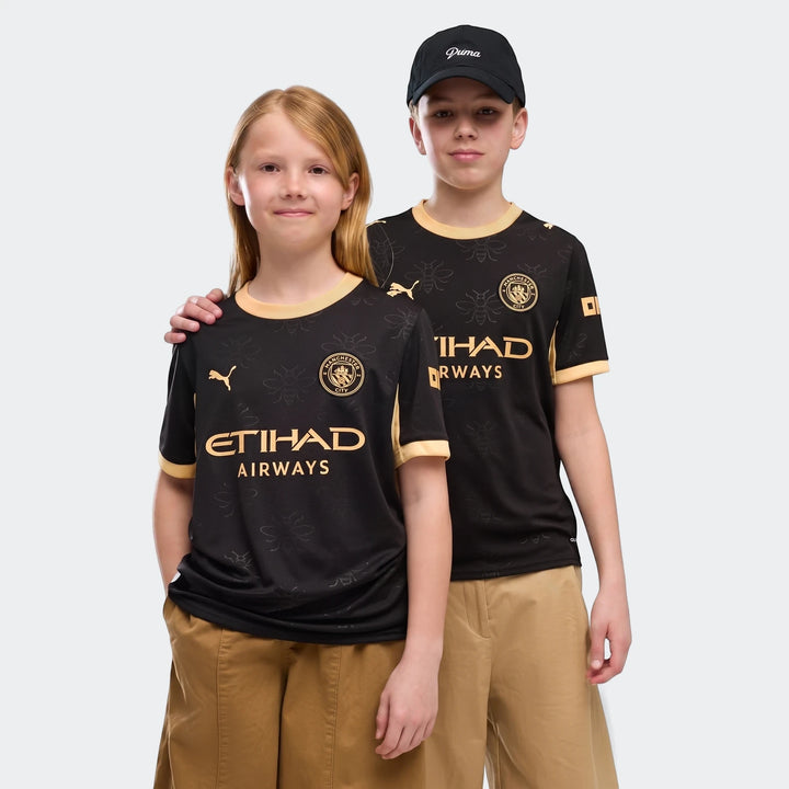 Two children model the black and gold Manchester City 26/27 Away Youth Jersey, featuring club crests and sponsor logos.