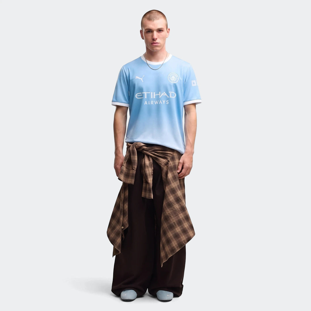 Male model showcasing the light blue Manchester City 26/27 Home Jersey, paired with dark pants and a plaid shirt.