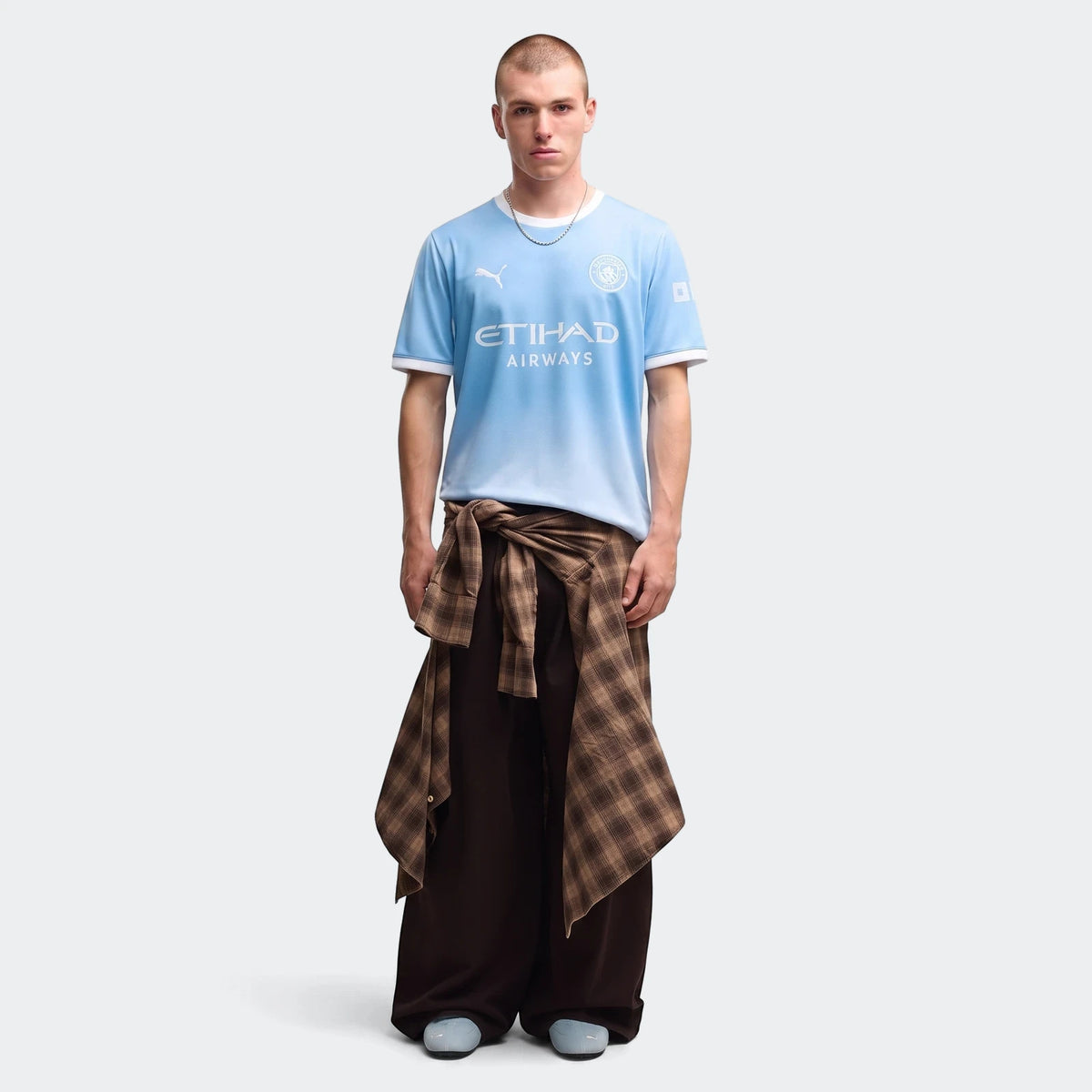 Male model showcasing the light blue Manchester City 26/27 Home Jersey, paired with dark pants and a plaid shirt.