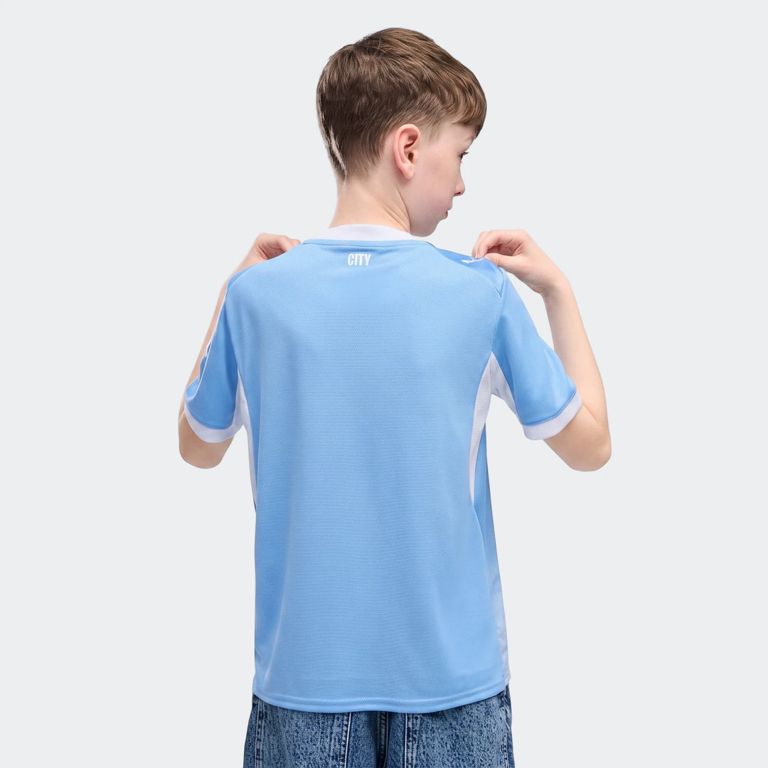 A young boy, seen from behind, wears a light blue and white Manchester City 26/27 Home Youth Jersey.