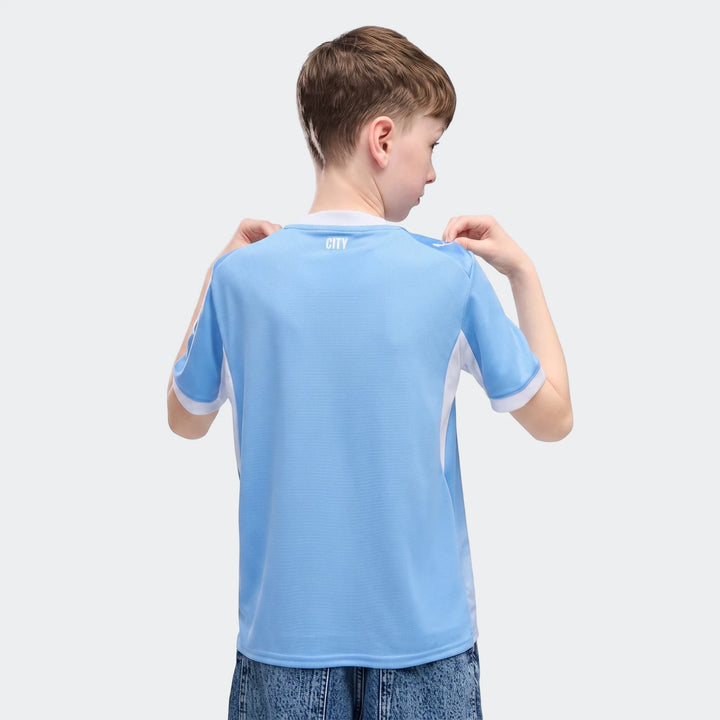 A young boy, seen from behind, wears a light blue and white Manchester City 26/27 Home Youth Jersey.