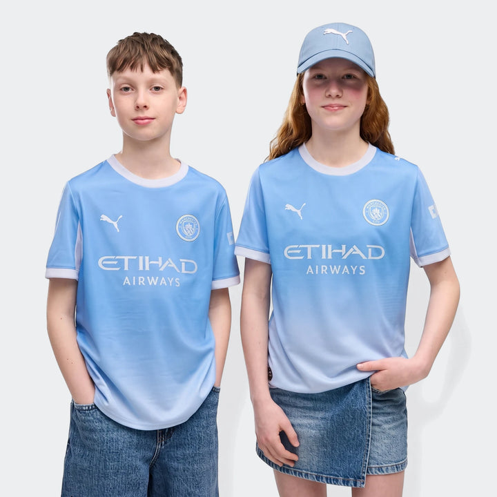 Youth boy and girl wearing light blue Manchester City 26/27 Home Jerseys with an ombre design.