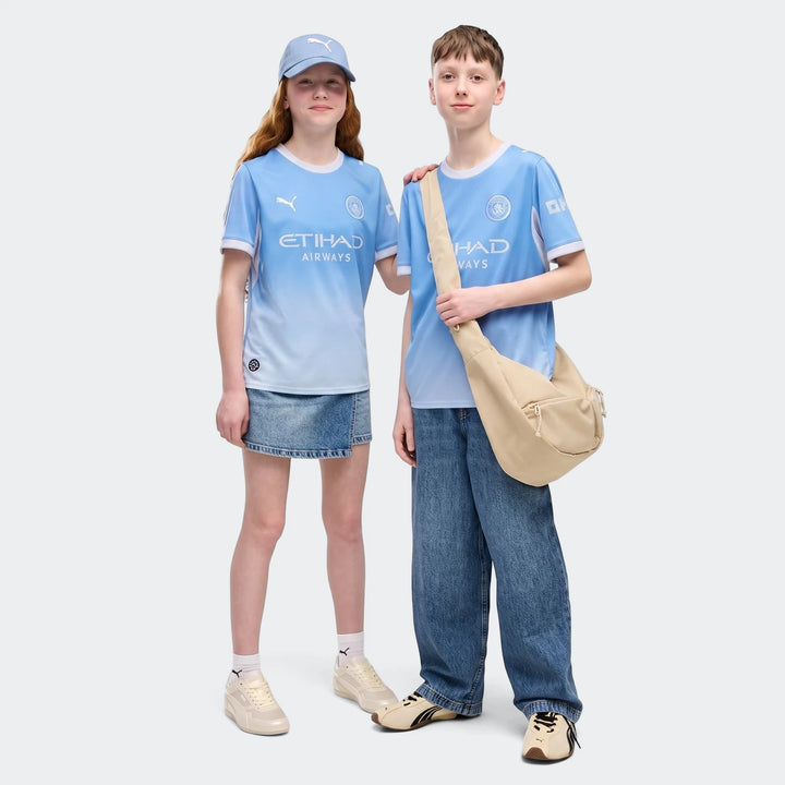 Two children model the Manchester City 26/27 Home Jersey, light blue with white trim, worn with jeans and a skirt.