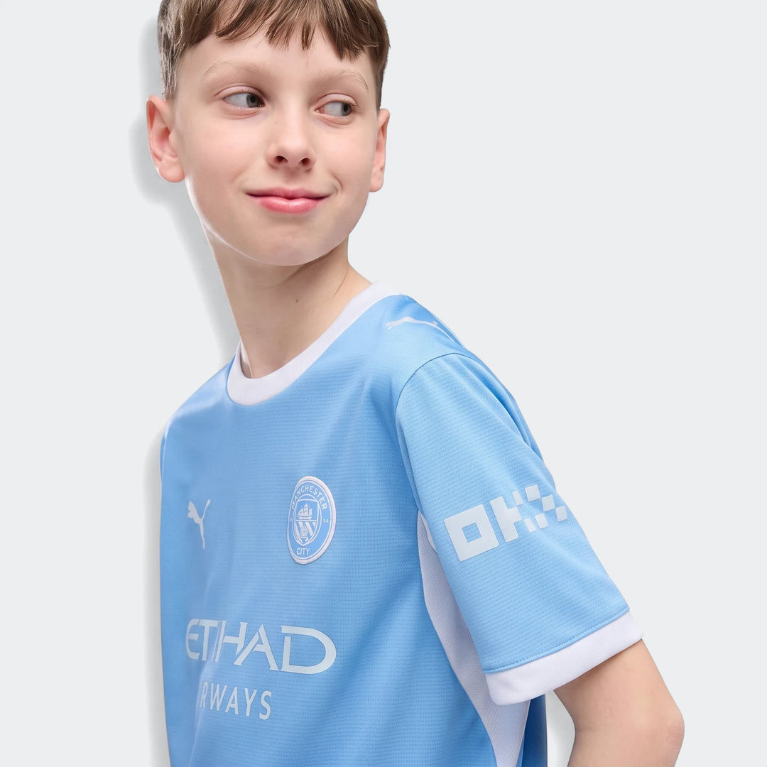 Young boy smiling in a light blue Manchester City 26/27 Home Jersey Youth, featuring club crest and sponsor logos.