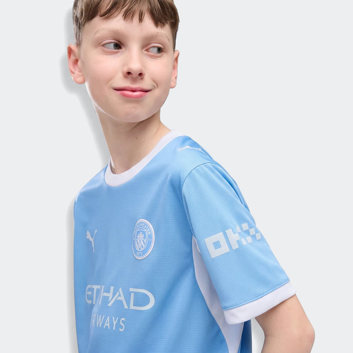 Young boy smiling in a light blue Manchester City 26/27 Home Jersey Youth, featuring club crest and sponsor logos.