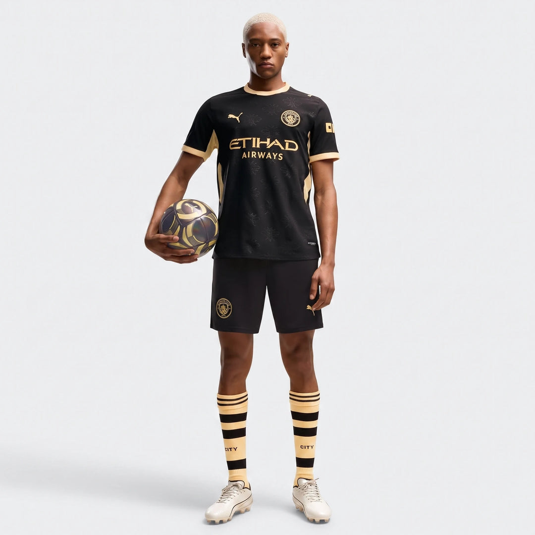 Male model wearing a Manchester City Away Authentic Jersey in black with gold accents, full kit, holding a soccer ball.