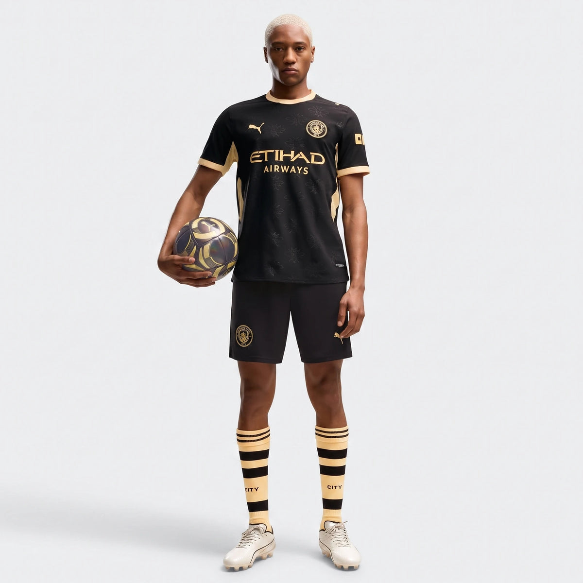 Male model wearing a Manchester City Away Authentic Jersey in black with gold accents, full kit, holding a soccer ball.