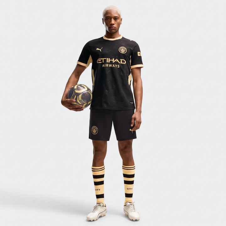 Male model wearing a Manchester City Away Authentic Jersey in black with gold accents, full kit, holding a soccer ball.