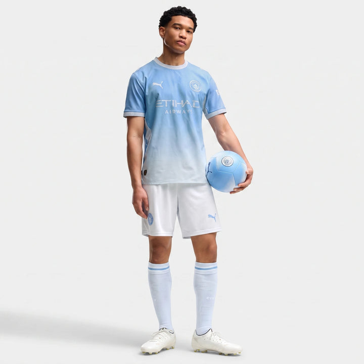 A man models the Manchester City Home Authentic Jersey for men, featuring a light blue to white gradient, white shorts, and