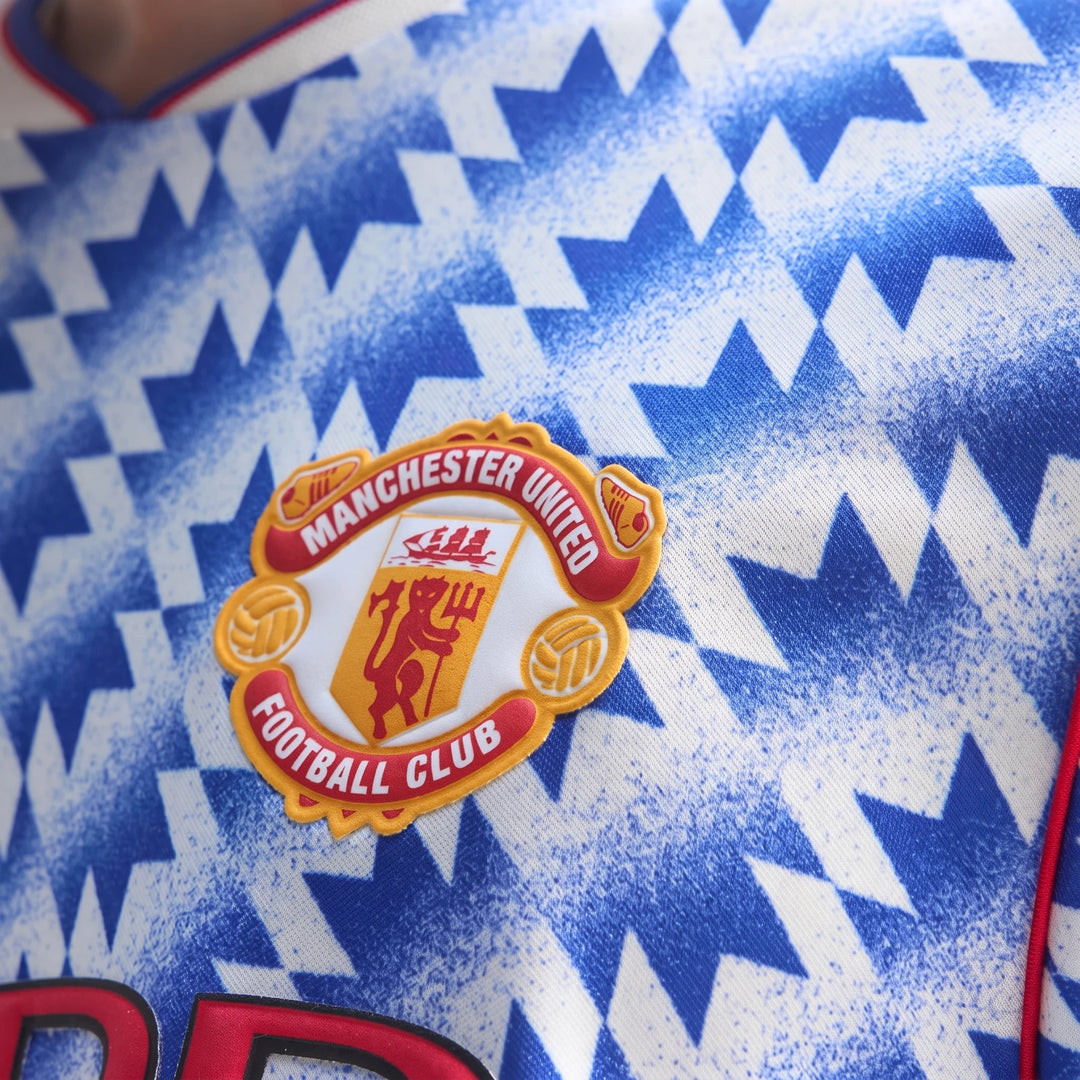 Close-up of Manchester United 1990 Away Jersey showing embroidered club crest on blue and white geometric fabric.