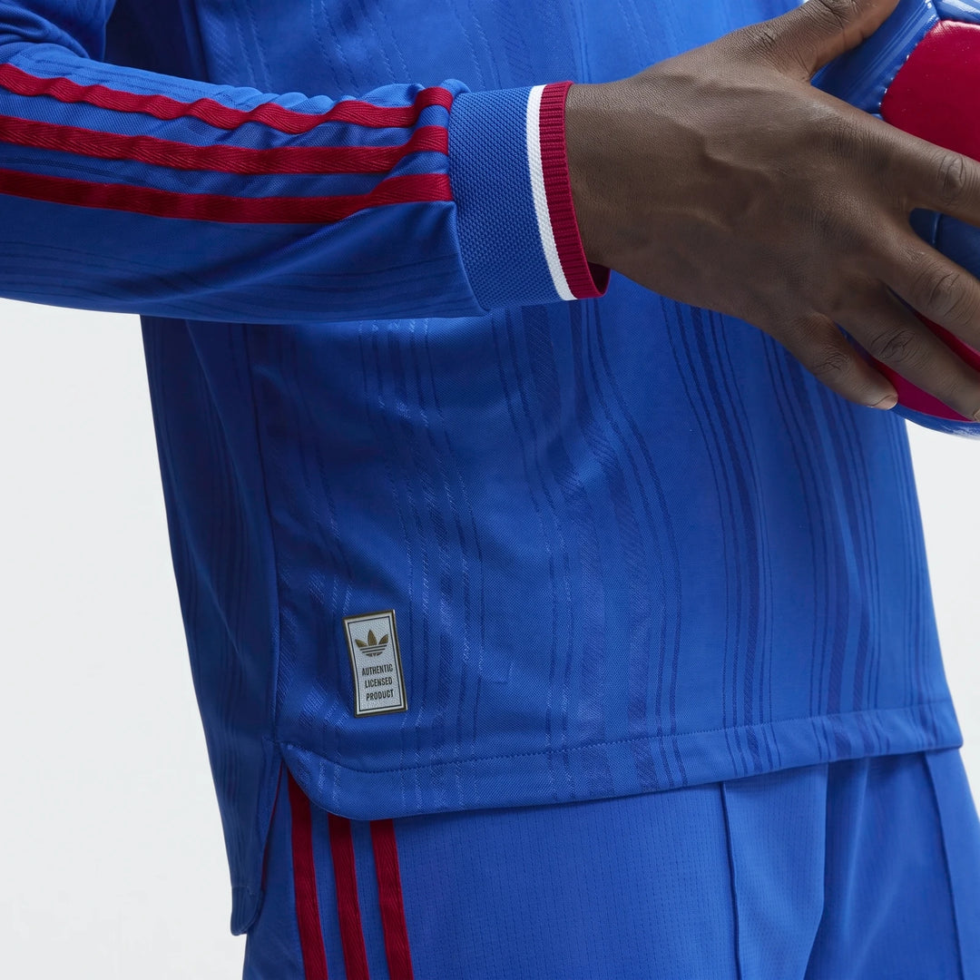 Close-up of a person wearing a blue Manchester United 26/27 Away Authentic Longsleeve Jersey with red stripes, holding a red