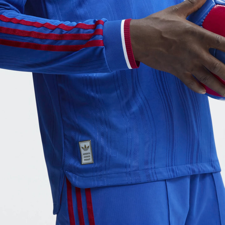 Close-up of a person wearing a blue Manchester United 26/27 Away Authentic Longsleeve Jersey with red stripes, holding a red