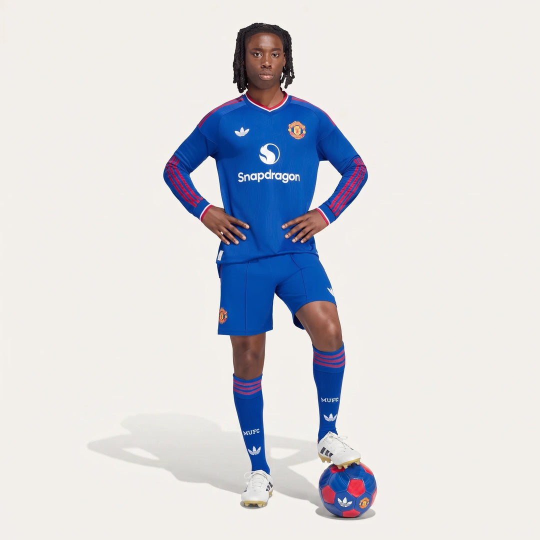 Male model in blue Manchester United 26/27 Away Authentic Longsleeve Jersey, matching shorts and socks, with foot on soccer