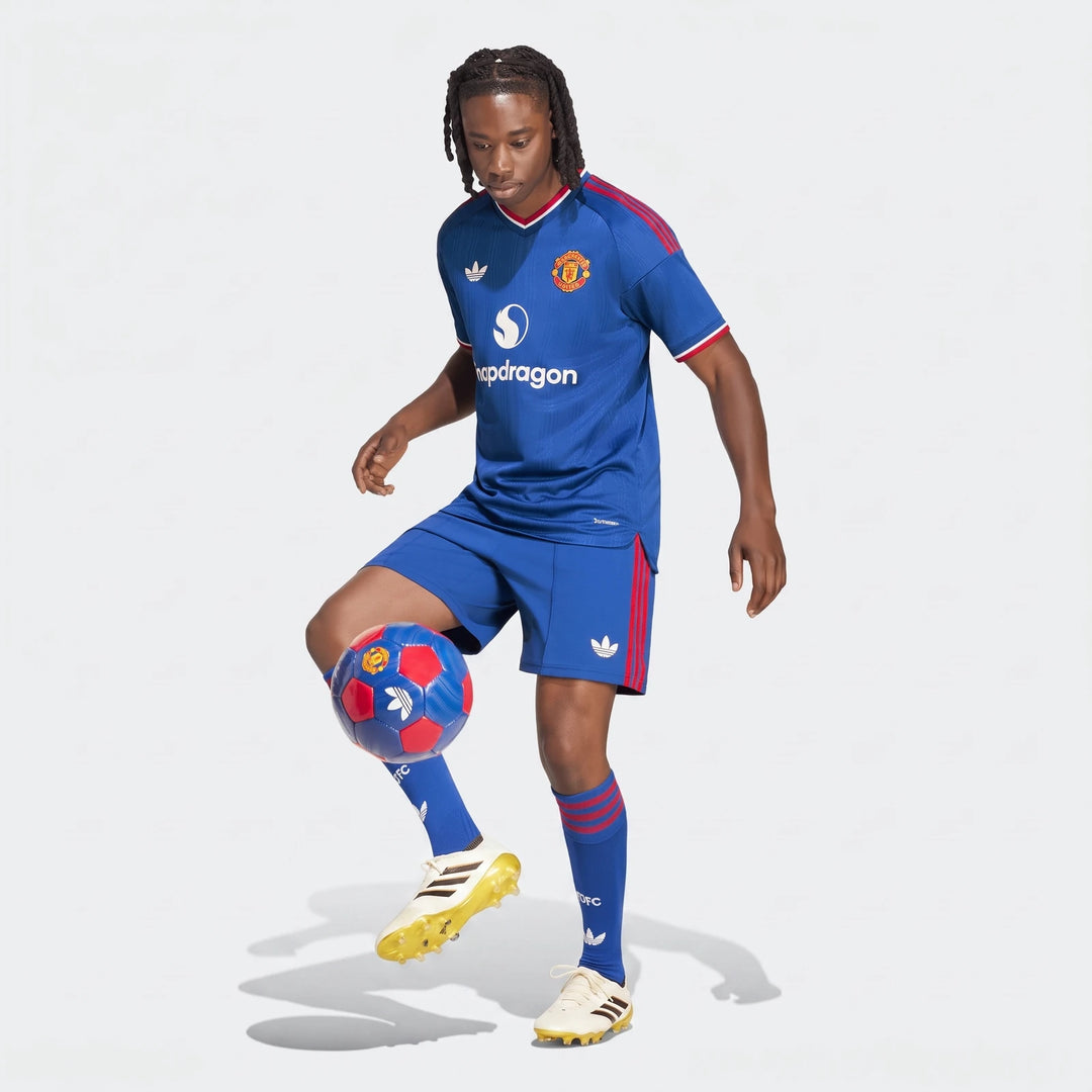 A young male model wearing the blue Manchester United 26/27 Away Authentic Jersey with red stripes, matching shorts, and