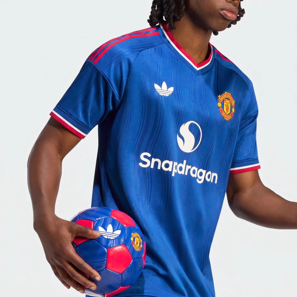 Model wearing a blue Manchester United 26/27 Away Jersey Authentic, featuring Adidas and Snapdragon logos, holding a soccer