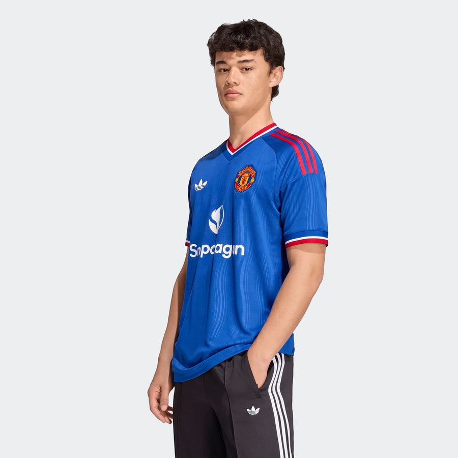 Male model wearing a blue Manchester United 26/27 Away Jersey with red shoulder stripes and Adidas branding, paired with