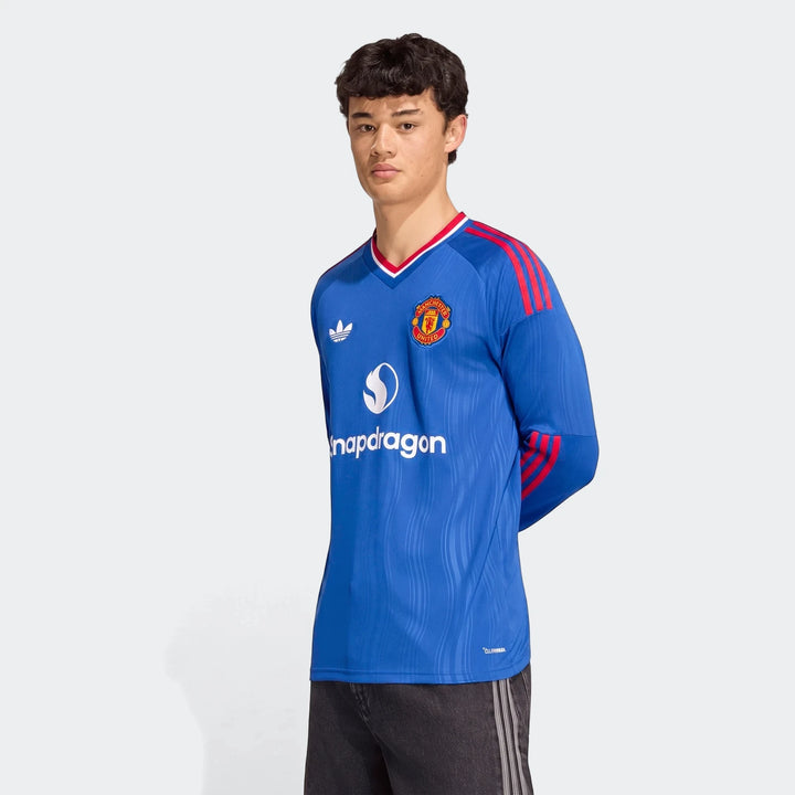 Young man models a blue long-sleeve Manchester United 26/27 away jersey with red stripes and sponsor logos.