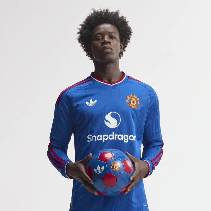 Young man wearing a blue Manchester United 26/27 Away Authentic Longsleeve Jersey with red stripes, holding a soccer ball.