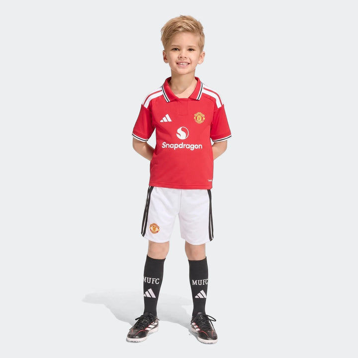 Young boy models the Manchester United 26/27 Home Mini Kit, featuring a red jersey, white shorts, and black socks.