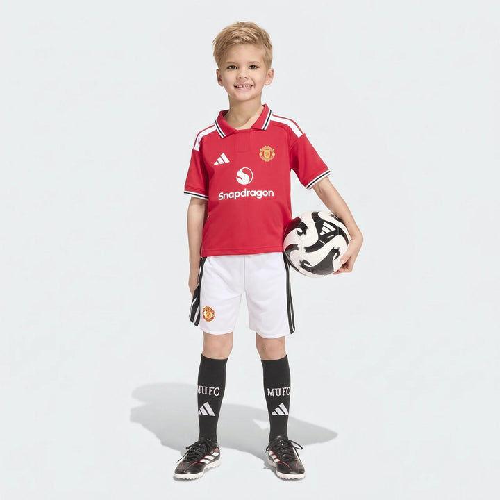 Young boy wearing a red and white Manchester United 26/27 Home Mini Kit, including jersey, shorts, and socks, holding a