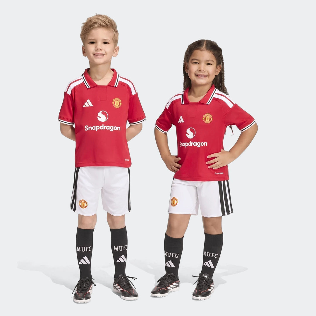 Two smiling children model the Manchester United 26/27 Home Mini Kit, featuring a red jersey, white shorts, and black socks,