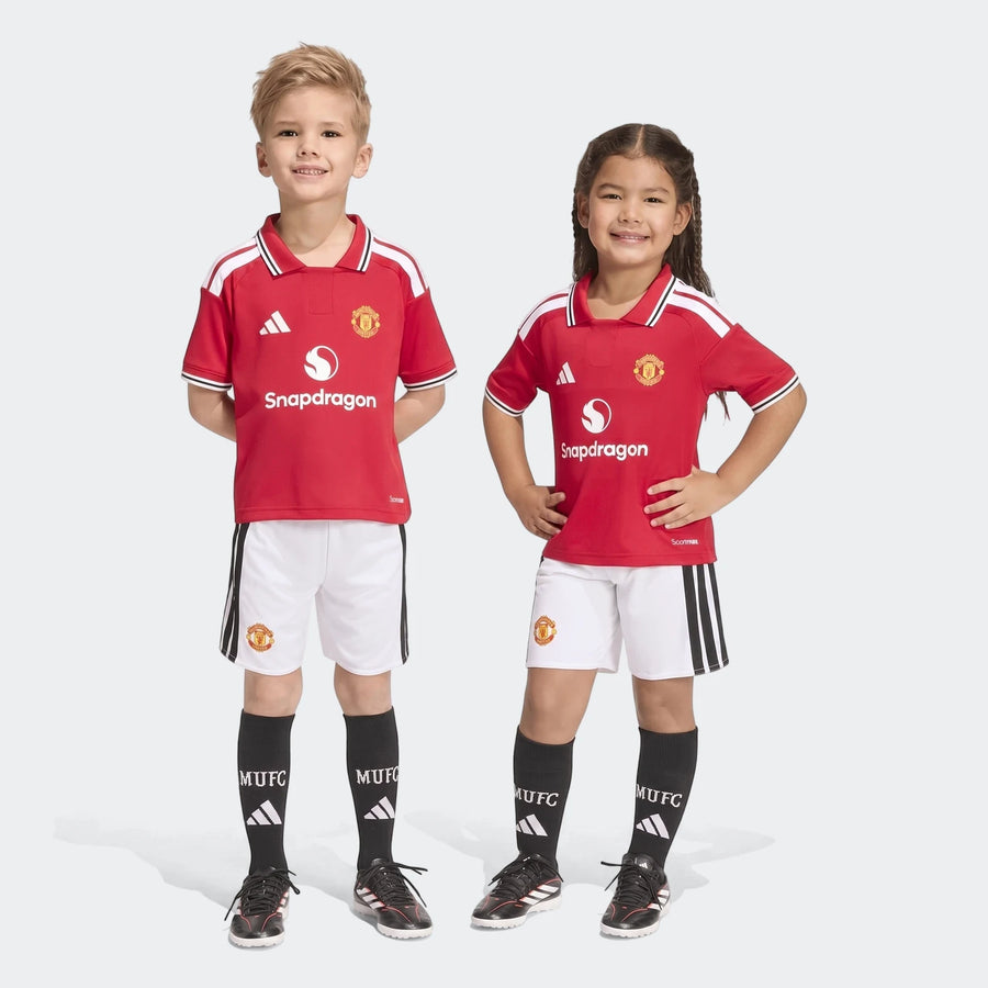 Two smiling children model the Manchester United 26/27 Home Mini Kit, featuring a red jersey, white shorts, and black socks,