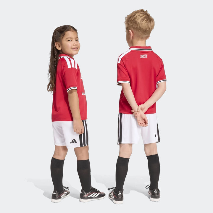 Two children model the Manchester United 26/27 Home Mini Kit, featuring a red jersey, white shorts, and black socks.