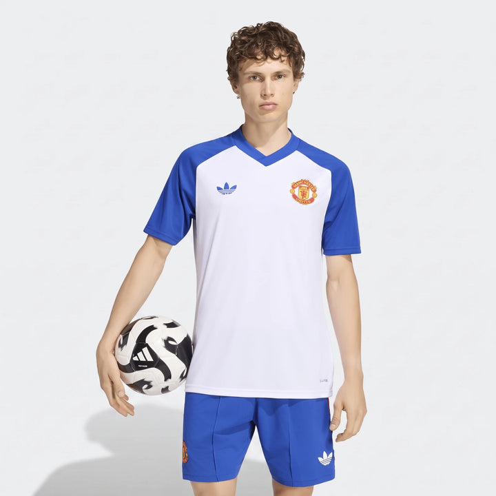 A male model wears the Manchester United 26/27 Pre Match Jersey in white and blue, holding a soccer ball.