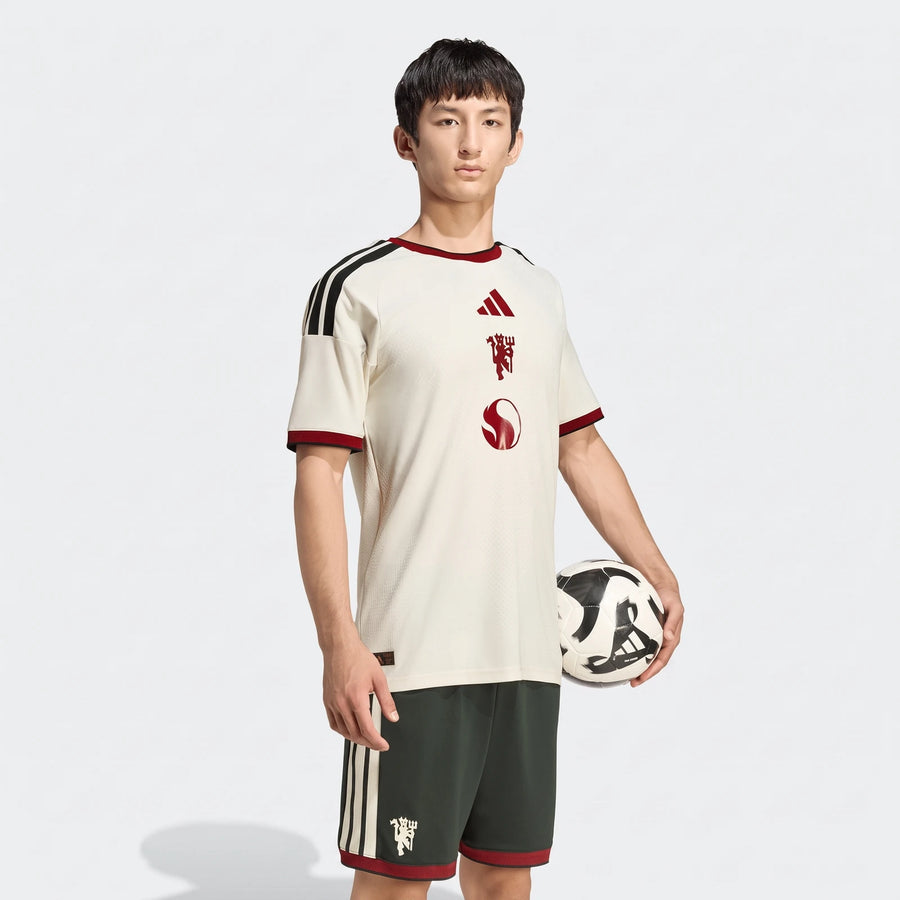 Male model wearing a cream Manchester United 26/27 Third Authentic Jersey with dark green shorts, holding a soccer ball.