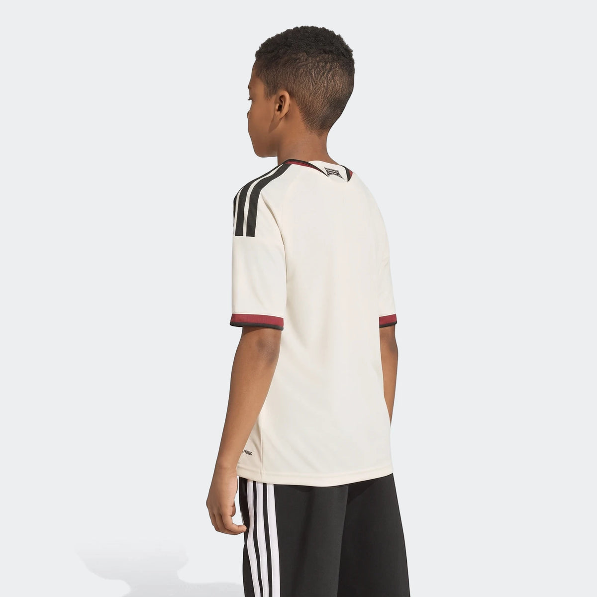 A young boy models the Manchester United 26/27 Third Jersey in cream with black stripes and red trim, viewed from the back.