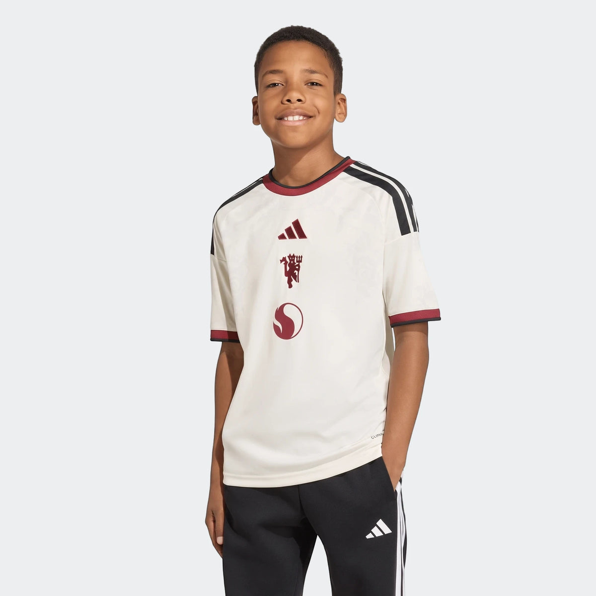Young boy wearing a cream/off-white Manchester United 26/27 Third Jersey with red and black details, standing with black