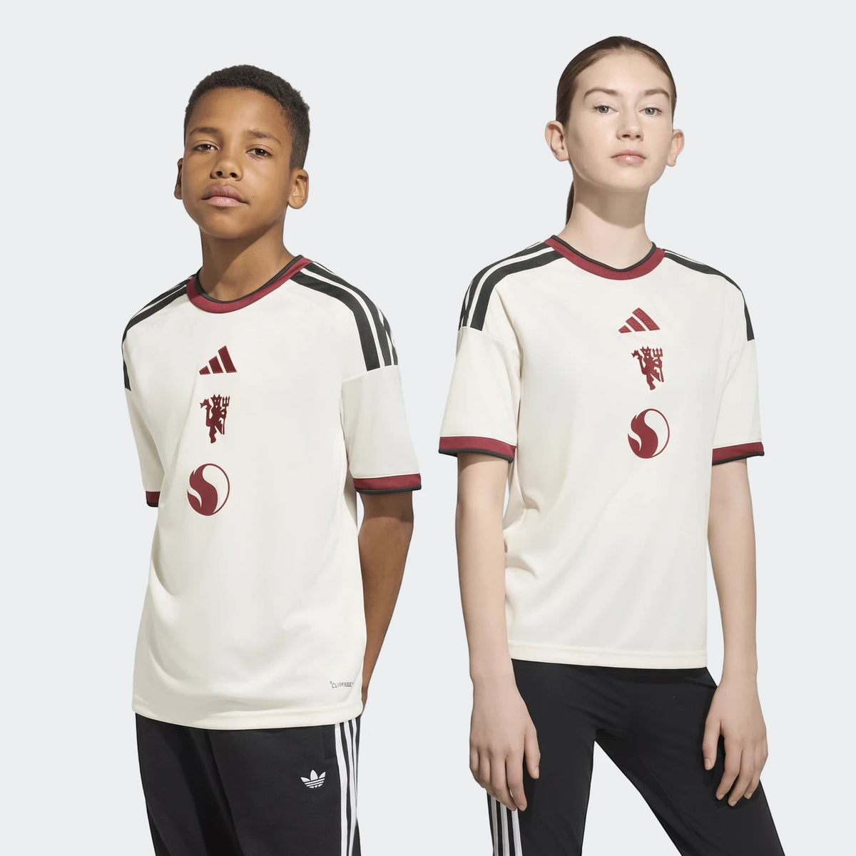 Two kids, a boy and a girl, model the Manchester United 26/27 Third Jersey, featuring an off-white design with black stripes