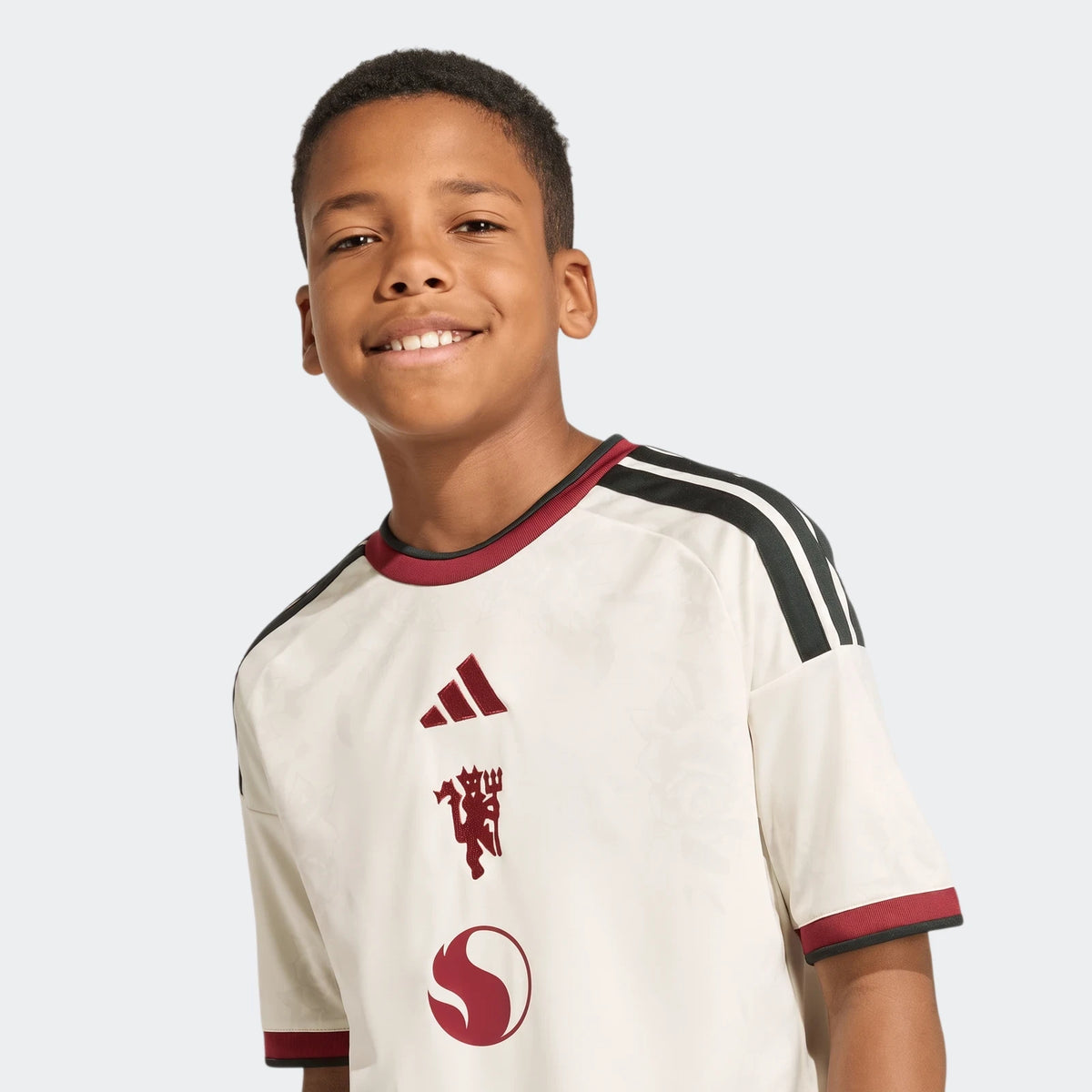 Smiling boy wearing an off-white, dark red, and black Adidas Manchester United 26/27 Third Jersey.