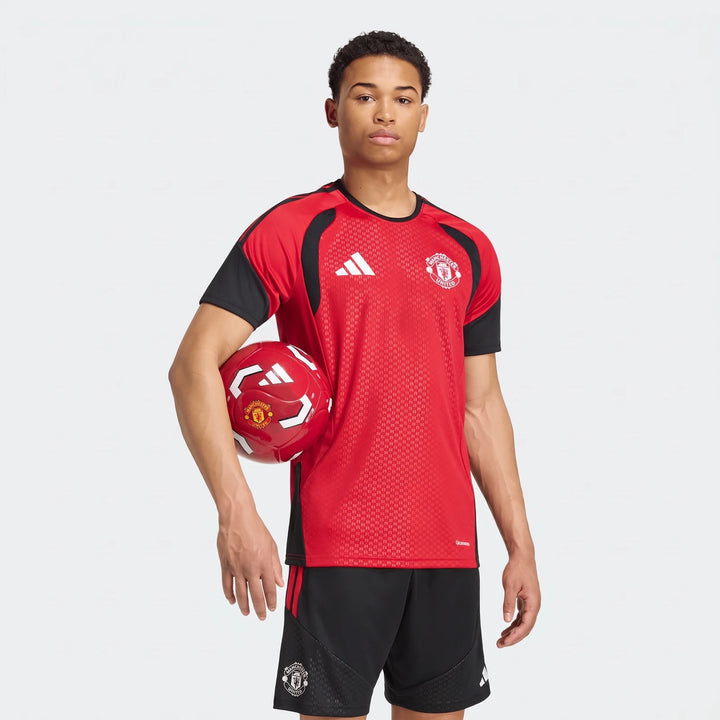 Young male model wearing a red and black Manchester United 26/27 Tiro Training Jersey and black shorts, holding a red soccer