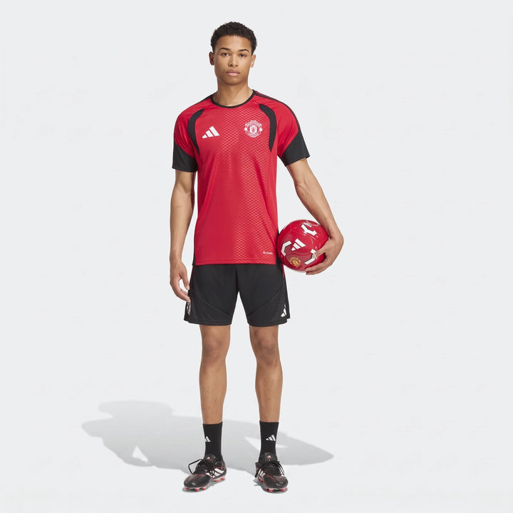 Male model wearing a red and black Manchester United 26/27 Tiro Training Jersey and black shorts, holding a soccer ball.
